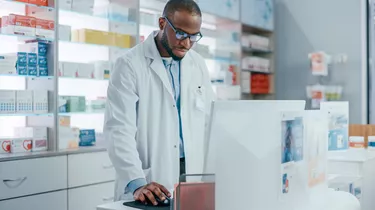 Pharmacy Drugstore Checkout Counter: Portrait of Professional Black Pharmacist Working on Computer, Checks Inventory of Medicine, Supply of Supplements, Health Care and Beauty Products, Vitamins