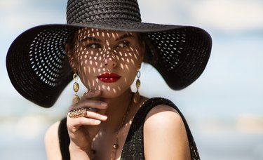 Woman in Hat Portrait. Fashion Luxury Model in Black Summer Hat with Make up and Golden Jewelry. Close up Beauty Face over Sky Background