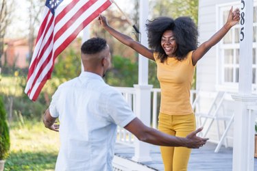 Smiling afro couple with wide open arms for big hug   in front their new house with the american flag on the porch