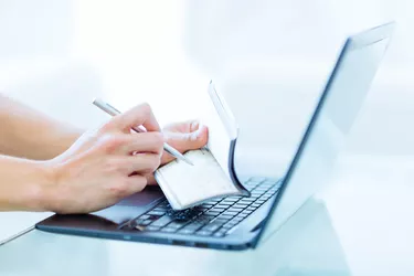 Persons hands writing a personal bank cheque using a pen while working on a laptop computer.