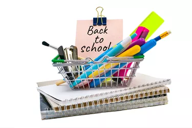 A shopping cart with office supplies on exercise books and words back to school on paper sticky sheets. Purchases for study, education concept on white background. Mini basket with stationery.
