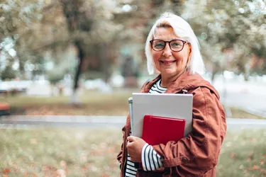 Adult caucasian blonde woman in glasses for sight with books, notebook and magazines looking at the camera walking in the autumn park