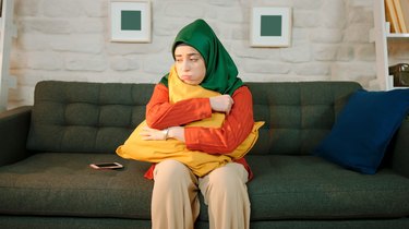Unhappy young woman sitting on sofa at home wearing a turban. The woman is hugging the pillow.