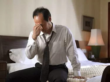 Businessman sitting on side of bed, looking tired.