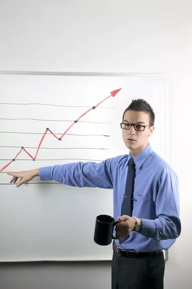 A businessman gives a presentation, gesturing to an upward pointing graph and holding his coffee mug as he speaks