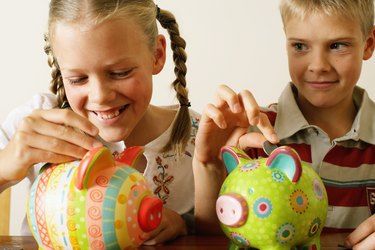 Twin brother and sister (10-12) dropping coins into piggy banks, girl laughing, close-up
