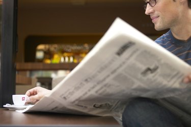 "Man sitting at cafe table reading newspaper, reaching for cup"