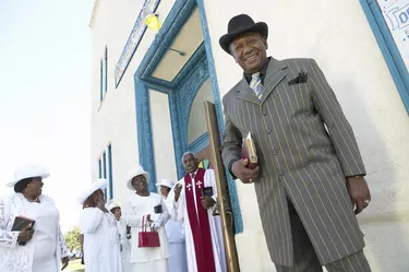 Well Dressed Man Outside a Church Standing and Looking at the Camera, with the Priest and Members of the Congregation in the Background