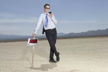 Businessman in dry lake bed leaning on 'for sale' sign