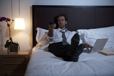 Businessman lying on bed using laptop and remote control