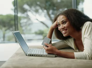 Young woman lying on bed with holding credit card and laptop, smiling, side view