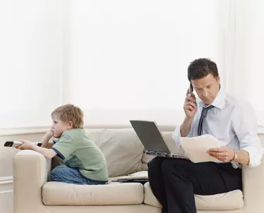 Businessman Sitting on a Sofa at Home With a Laptop and Using a Mobile Phone and His Son Watching TV