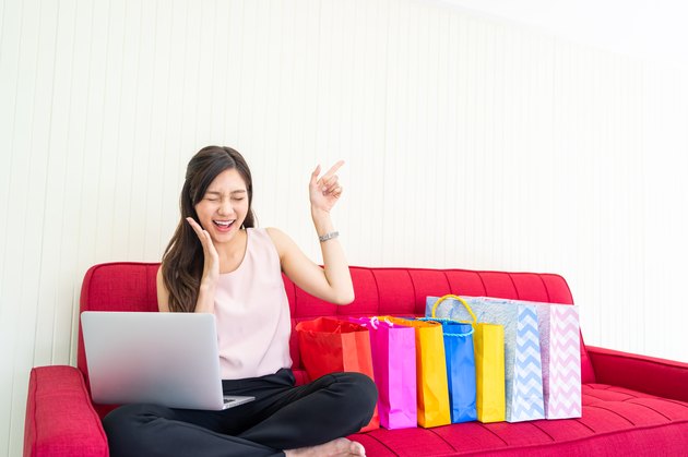 Portrait of young woman using laptop for online shopping on sofa and pointing fingers to empty space.