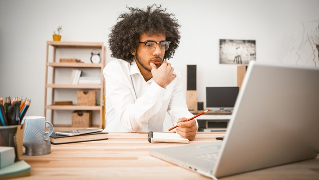 Pensive young man is engaged at home in front of computer. Arab student or businessman taking notes in a notebook. Home education concept. Toned image