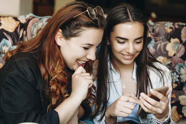 Beautiful plus size women with red hair looking at a smartphone screen smiling holded by her girlfriend while sitting in coffee shop.