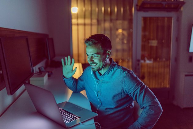 Smiling Caucasian freelancer waving and having video call with his boss over laptop. In ears earphones. Late night work concept.