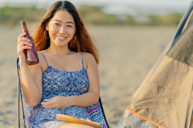 Portrait of young and beautiful woman sitting on outdoor chair and holding beer and smiling happily for camera