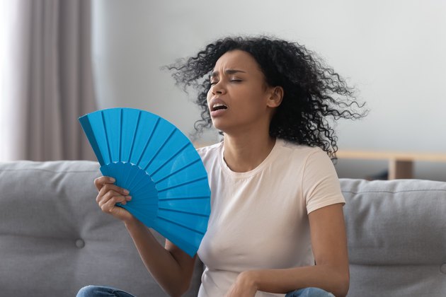 Overheated african young woman feeling hot waving fan at home