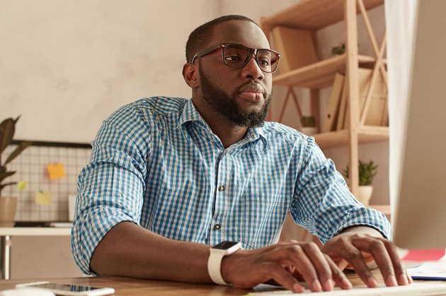 Busy day. Smart young afro american man in glasses working on the computer while sitting at his working place at home. Afro american businessman working remotely at home