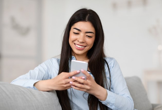 Smiling Asian Girl Browsing Social Networks On Smartphone At Home, Enjoying Weekend
