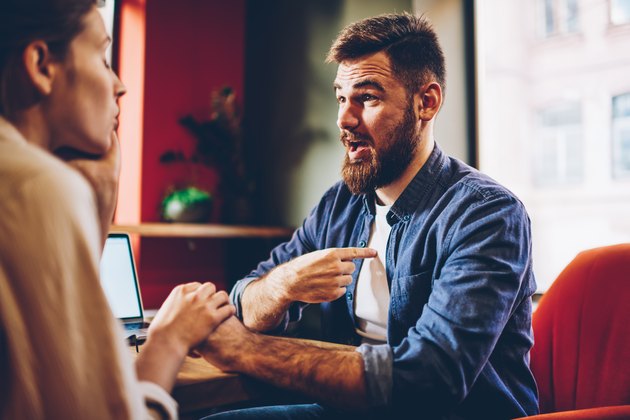 Wondered bearded young man solving conflict with girlfriend holding hands each other on romantic date.Surprised male discussing obligations with wife during quarrel sitting in restaurant interior