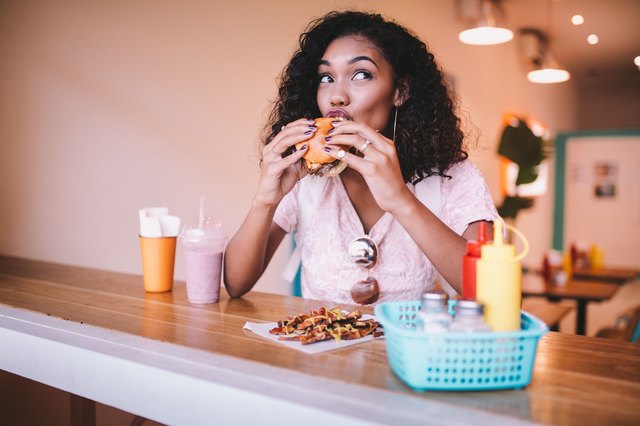 Young Black woman eating cheeseburger in diner with impish expression
