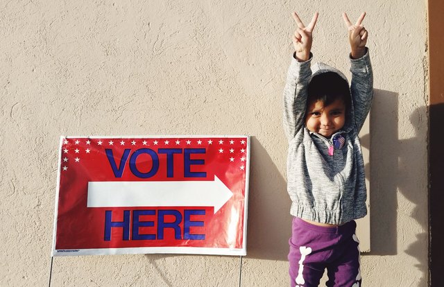 Small child flashing peace signs next to "Vote Here" sign