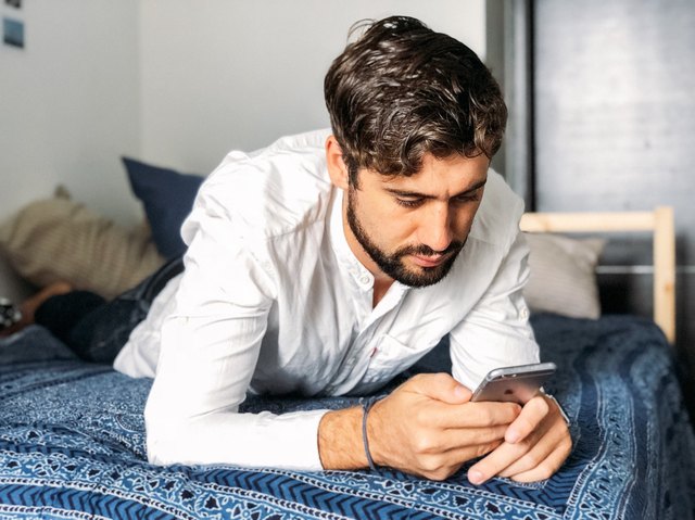 Young bearded man checking phone lying on his stomach on bed
