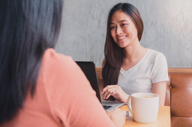 Young East Asian woman on laptop smiling at another woman over coffee