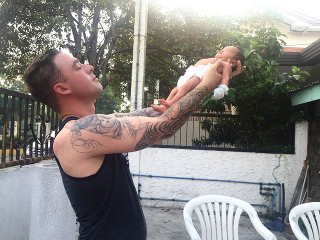 Tattooed young father holding newborn aloft on outdoor patio