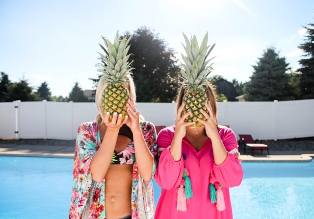 Two women by a pool holding pineapples in front of their faces