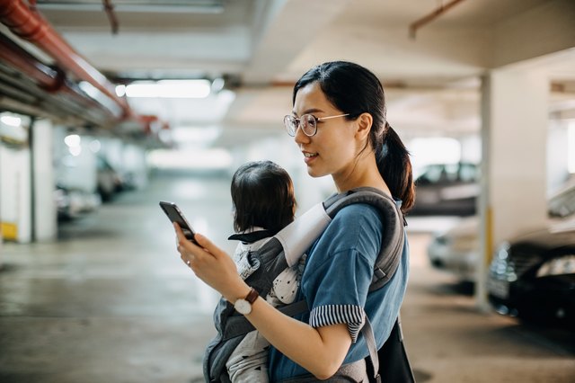 Busy young woman with baby in carrier using smartphone while walking to her car in car park