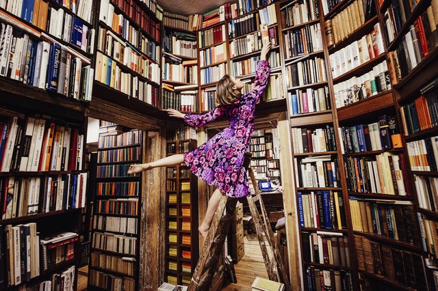 Carefree woman on ladder reaching for book in library