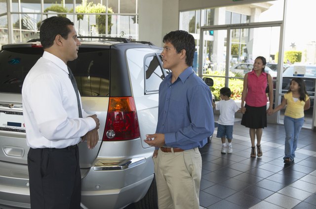 Man shaking car salesman's hand, family in background