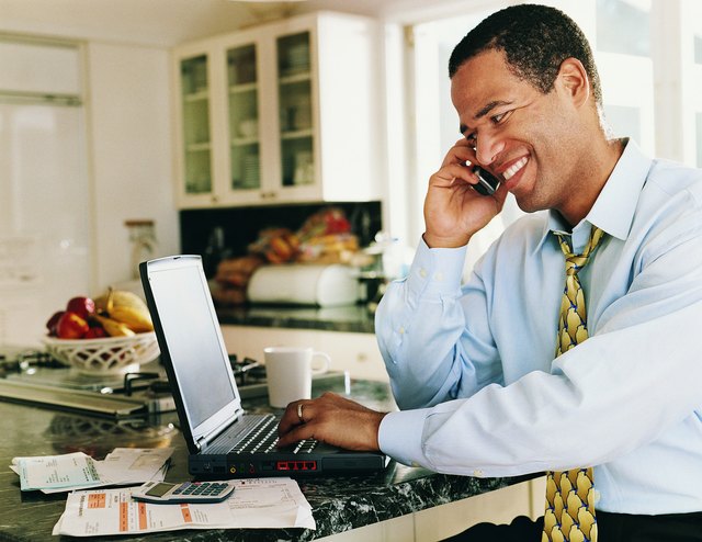 Man Sitting in a Kitchen Typing into a Laptop and Listening to his Mobile Phone